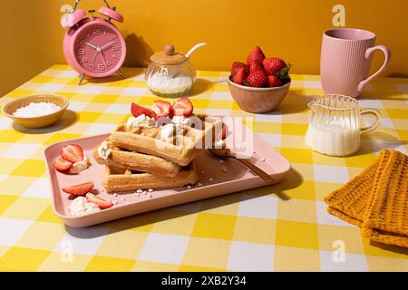 Une scène de petit déjeuner lumineuse et joyeuse avec gaufres dorées garnies de crème et de fraises fraîches sur une assiette rose, accompagnées d'ingredie Banque D'Images