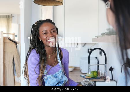 Une jeune femme joyeuse aux cheveux tressés, portant une chemise lavande et une salopette en denim, rit joyeusement dans une cuisine moderne et lumineuse Banque D'Images