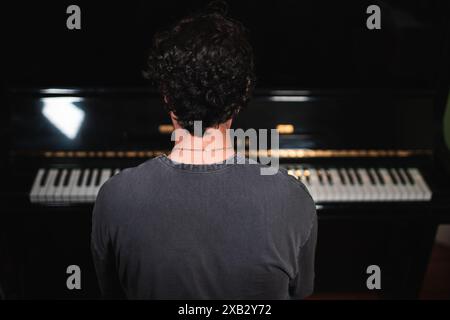 Une vue arrière d'un homme debout contemplativement devant un piano à queue dans une pièce légèrement éclairée, créant une scène d'anticipation et de réflexion artistique Banque D'Images