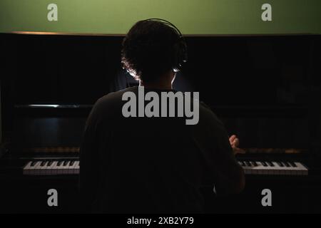 Un homme dans une pièce sombre avec un piano à queue, vu de derrière, porte des écouteurs tout en engageant intensément avec la musique L'image capture un moment d'artisti Banque D'Images