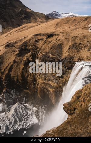 Une cascade majestueuse descend en cascade des falaises escarpées dans un abîme brumeux, sur fond de montagnes enneigées et de collines herbeuses sèches de l'Islande sce Banque D'Images