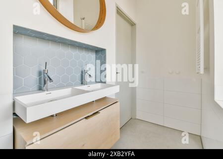 Cette image présente un intérieur de salle de bains contemporain avec un meuble-lavabo en bois avec un double évier blanc et élégants carreaux de dosseret hexagonaux. Banque D'Images