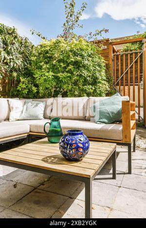 Un patio extérieur accueillant avec un canapé d'angle moderne, une table basse en bois et des vases décoratifs en verre bleu, entouré d'une végétation luxuriante. Banque D'Images