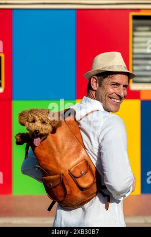 Un homme souriant portant un chapeau porte un Caniche jouet dans un sac à dos, posant devant un mur multicolore vibrant Banque D'Images