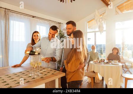 Un jeune homme souriant verse du champagne dans des verres dans la salle à manger de la maison. Les couples mariés de différents âges célèbrent le réveillon de Noël. Un gros hap Banque D'Images