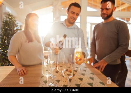 Le jeune homme souriant verse du champagne dans des verres dans la salle à manger de la maison. Deux couples mariés attirants célèbrent la veille de Noël. Heureux amis ce Banque D'Images