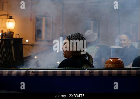 Femme avec un foulard à motifs travaillant à l'intérieur d'un stand de nourriture pendant les festivités de l'alfama, entourée de fumée, des collègues en arrière-plan Banque D'Images