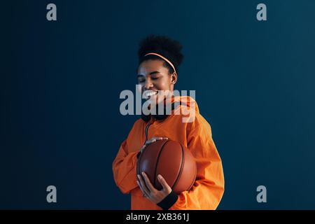 Femme souriante avec un ballon de basket portant des vêtements de sport orange et des écouteurs sans fil. Jeune femme aux cheveux bouclés tenant un ballon de basket. Banque D'Images