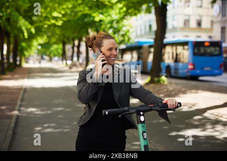 Femme d'affaires mature souriante avec scooter électrique poussant parler sur le téléphone portable sur le sentier Banque D'Images