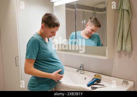 Jeune femme enceinte touchant l'abdomen par la tondeuse à cheveux devant le miroir à la salle de bain Banque D'Images
