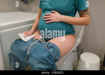 Section médiane d'une jeune femme enceinte touchant l'abdomen assise sur le siège des toilettes Banque D'Images
