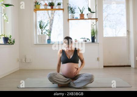Jeune femme enceinte touchant l'abdomen tout en étant assise sur un tapis d'exercice à la maison Banque D'Images