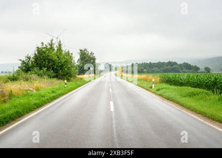 Route déserte à travers la campagne par une journée d'été brumeuse. Les éoliennes sont visibles à distance. Banque D'Images