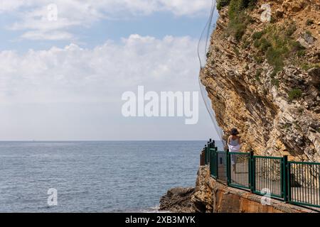 Une femme en robe blanche marche le long d'un sentier côtier près de falaises abruptes, regardant vers la mer. Le ciel est partiellement couvert de nuages, créant une scène pittoresque. Copier l'espace. Banque D'Images