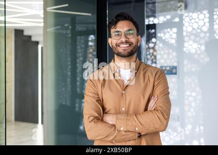 Portrait d'un jeune indien portant des lunettes et une chemise brune debout dans un bureau moderne, croisant ses bras au-dessus de sa poitrine et regardant avec confiance la caméra. Banque D'Images