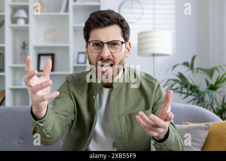 Portrait en gros plan d'un jeune homme en colère et inquiet assis sur un canapé à la maison, parlant lors d'un appel vidéo et criant émotionnellement à la caméra, faisant des gestes avec les mains. Banque D'Images