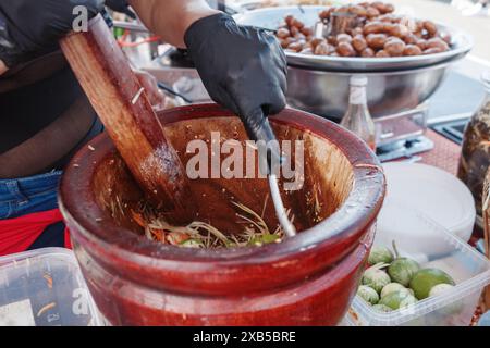 Vue rapprochée d'une personne portant des gants noirs en utilisant un mortier en bois et un pilon pour préparer une salade traditionnelle de papaye thaïlandaise, également connue sous le nom de Som Tum. Banque D'Images