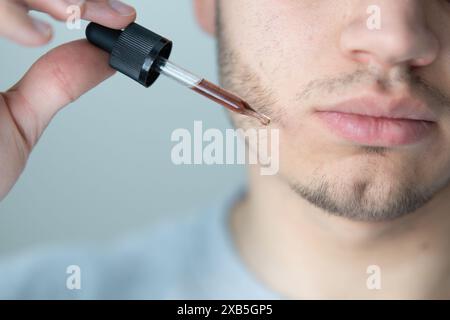 jeune homme appliquant l'huile de croissance de barbe avec pipette gros plan. homme tenant la pipette avec de l'huile pour la barbe, appliquant le sérum cosmétique avec la pipette sur le visage Banque D'Images