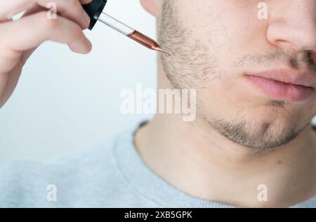 jeune homme appliquant l'huile de croissance de barbe avec pipette gros plan. homme tenant la pipette avec de l'huile pour la barbe, appliquant le sérum cosmétique avec la pipette sur le visage Banque D'Images