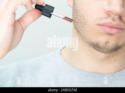 jeune homme appliquant l'huile de croissance de barbe avec pipette gros plan. homme tenant la pipette avec de l'huile pour la barbe, appliquant le sérum cosmétique avec la pipette sur le visage Banque D'Images