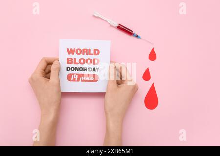 Mains féminines tenant une feuille de papier avec signe WORLD BLOOD DONOR DAY 14 JUIN, gouttes de sang en papier et seringue sur fond rose Banque D'Images