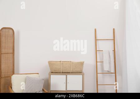 Fauteuil avec coussin, panier en osier sur table de chevet et échelle en bois dans la chambre Banque D'Images