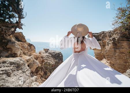 Une femme en robe blanche se tient debout sur une falaise rocheuse surplombant l'océan. Elle porte un chapeau de paille et elle profite de la vue Banque D'Images