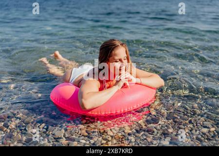 Une femme est allongée sur un radeau gonflable rose dans l'océan. Elle porte un bikini blanc et a les mains sur les hanches. La scène est paisible et détendue Banque D'Images