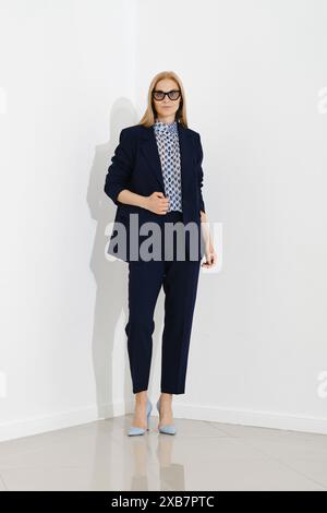 Femme professionnelle se tient en toute confiance contre un mur blanc, vêtue d'un costume d'affaires bleu marine chic associé à des talons bleu clair Banque D'Images