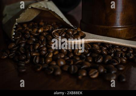 Grains de café fraîchement torréfiés se répandant d'un sac en papier brun partiellement ouvert sur une surface en bois. Une cuillère en bois se trouve parmi les grains Banque D'Images