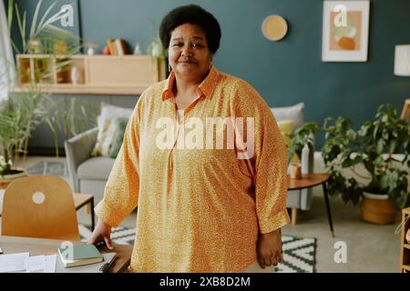 Femme d'affaires afro-américaine mature debout à côté du bureau de travail tout en travaillant à distance et souriant avec le sourire fatigué Banque D'Images