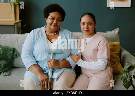 Jeune infirmière tenant soigneusement la main des femmes afro-américaines matures pendant qu'elles sont assises sur le canapé et posent pour la photo Banque D'Images