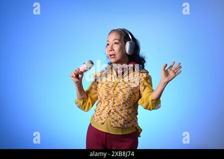 Élégante dame d'âge moyen dans le casque chantant avec microphone sur fond bleu Banque D'Images