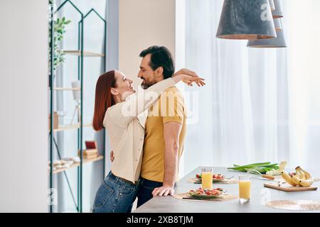 Un homme barbu et une femme rousse partageant un câlin chaleureux dans une cuisine moderne, créant un moment d'intimité et de convivialité. Banque D'Images