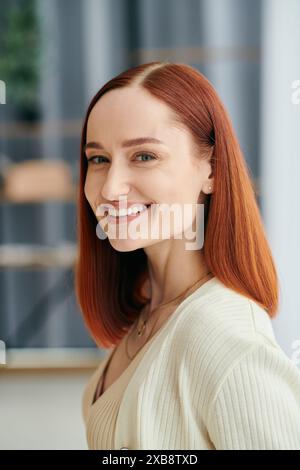 Une femme aux cheveux cramoisis souriant chaleureusement à la caméra, exsudant bonheur et confort dans un cadre d'appartement moderne. Banque D'Images