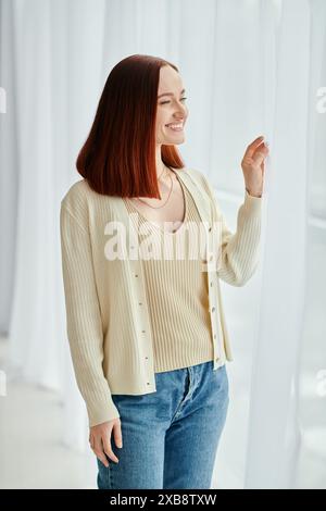 Une belle femme rousse portant un cardigan et un Jean se tient en toute confiance devant une fenêtre, dégageant une élégance décontractée. Banque D'Images