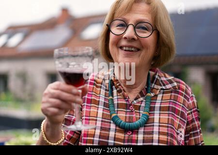 Une femme âgée sourit tout en tenant un verre de vin rouge, portant une chemise à carreaux et un collier vert, célébrant à la maison - heureuse dame senior toasting Banque D'Images