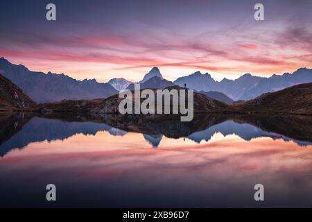 Beau et coloré ciel de feu au-dessus d'une chaîne de montagnes se reflétant dans un lac au coucher du soleil, avec une silhouette de randonneur au sommet d'une colline Banque D'Images