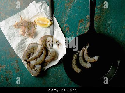 Crevettes fraîches préparées pour la cuisson sur une surface sarcelle. Les crevettes sont sur du papier blanc avec un couteau, un coin de citron, et certaines dans une poêle. Banque D'Images