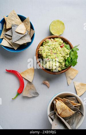 Un bol de guacamole frais garni de coriandre, entouré de chips de tortilla bleues et blanches, un piment rouge, une gousse d'ail et un citron vert coupé en deux Banque D'Images