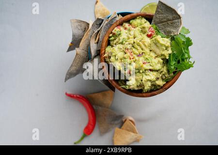 Un bol de guacamole frais garni de coriandre, entouré de chips de tortilla bleues et blanches, un piment rouge, une gousse d'ail et un citron vert coupé en deux Banque D'Images