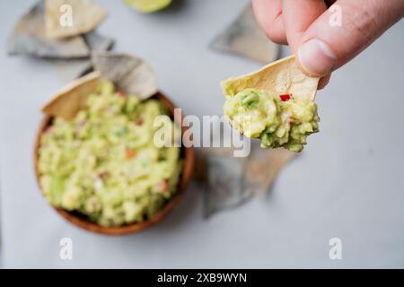 Main tenant une puce de tortilla au guacamole au-dessus d'un bol en bois rempli de guacamole, garni de coriandre. Coin de citron vert et chips tortilla de maïs bleu Banque D'Images