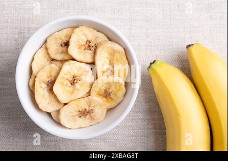 Chips de banane ou chips de banane dans un bol blanc sur tissu de lin. Tranches de bananes frites jaunes croquantes, recouvertes de sucre ou de miel. Banque D'Images