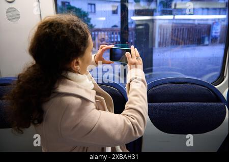 Une femme prend une photo à travers la fenêtre d'un train un jour de pluie. Elle utilise son smartphone pour documenter le paysage. Banque D'Images