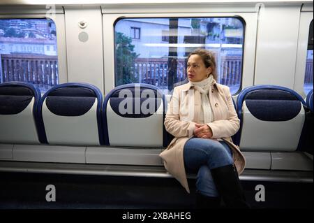 Une femme est assise seule sur un train portant un manteau beige et une écharpe. Elle regarde par la fenêtre le paysage urbain, véhiculant un sentiment de contemplation et de solitu Banque D'Images