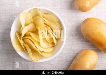 Chips de pommes de terre salées dans un bol blanc sur tissu de lin. Chips, fines tranches de pommes de terre frites, et à droite des pommes de terre crues. Banque D'Images