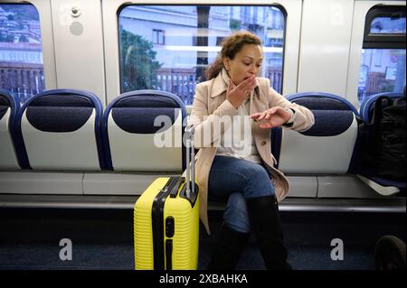 Femme assise sur un banc de train avec une valise jaune, regardant la montre avec une expression inquiète. Capturer les thèmes du voyage, du stress et de l'anxiété en public Banque D'Images