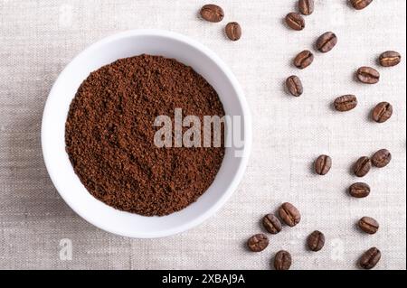 Café en poudre, grains de café fraîchement moulus dans un bol blanc sur tissu de lin. Sur la droite graines grillées de baies de Coffea arabica. Banque D'Images