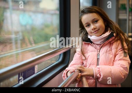 Une jeune fille portant un manteau d'hiver rose monte dans un train, regardant par la fenêtre avec une expression calme. Le paysage à l'extérieur de la fenêtre est flou. Banque D'Images