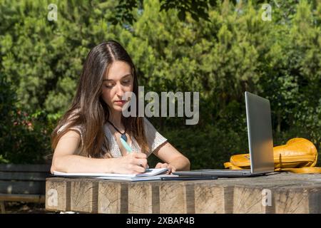 Jeune fille d'université assise sur le campus avec son ordinateur portable, étudiant un jour ensoleillé. Vie académique, concentration, technologie, préparation aux cours Banque D'Images